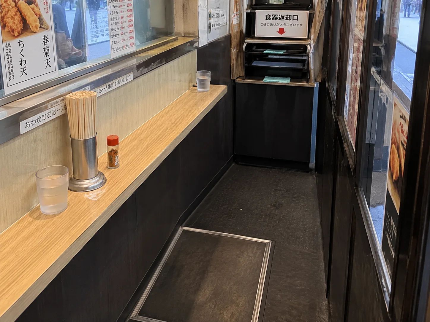 A narrow soba counter with chopsticks, condiments, water cups, and one empty standing space.