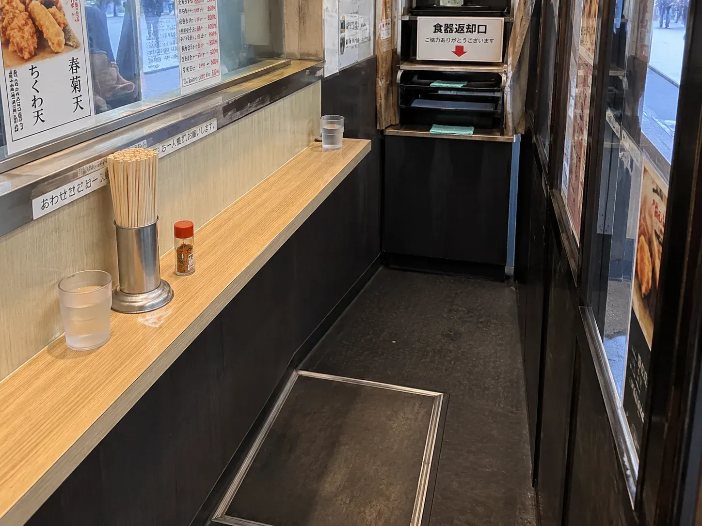 A narrow soba counter with chopsticks, condiments, water cups, and one empty standing space.